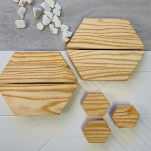 May include: Wooden hexagon-shaped decorative items. The image shows two large, split hexagons and three smaller ones, all with a natural wood grain pattern. White stones are scattered near the top. The items are on a white and gray surface.