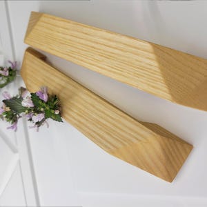 May include: Two wooden cabinet pulls with a light wood grain. The pulls have a geometric design with angled edges. A sprig of purple and green flowers rests near the pulls on a white surface.