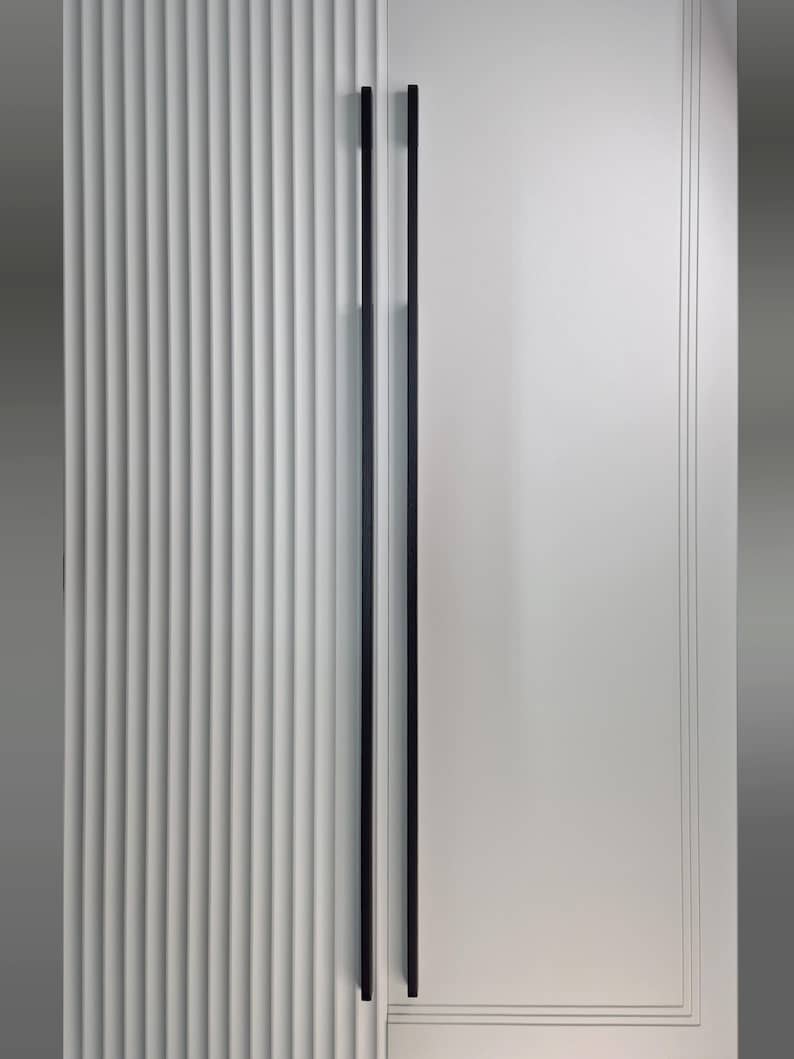 May include: A close-up of a white door with two long, black handles. The left side of the door has vertical, ridged detailing, while the right side is smooth with subtle linear accents. The handles are sleek and modern.
