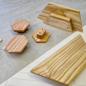 May include: A collection of unfinished wooden cabinet hardware, including hexagonal knobs and rectangular pulls. The natural wood grain is visible on each piece, with a light brown color. The hardware is arranged on a gray and white tiled surface.