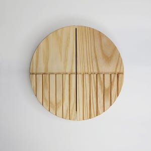 Pode incluir: Decoração de parede redonda em madeira clara. A metade superior é lisa, enquanto a metade inferior apresenta linhas verticais ranhuradas. A madeira tem um acabamento claro e natural, dividida em quatro seções.