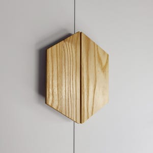 Pode incluir: Um puxador de armário de madeira em forma geométrica de hexágono. O puxador é feito de madeira clara com grão visível e é montado em uma superfície branca. O design é moderno e minimalista.