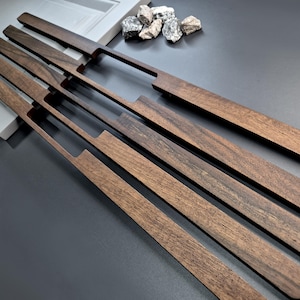 May include: Four long, rectangular dark brown wooden cabinet handles with a smooth finish and a recessed grip section on one side. The handles are arranged diagonally in a parallel row, showcasing the natural wood grain patterns and texture.