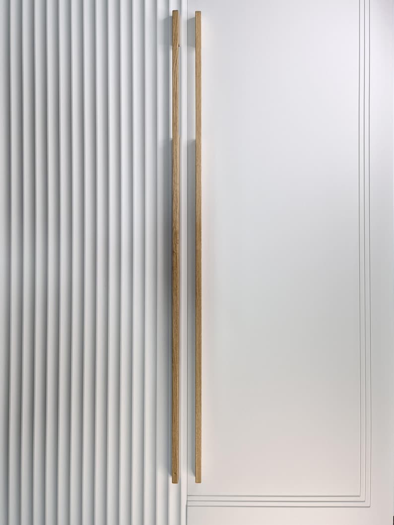 May include: A white door with vertical ribbed paneling on the left and a smooth white panel on the right. Two vertical wooden door pulls are mounted on the right panel. The door pulls are made of light-colored wood.