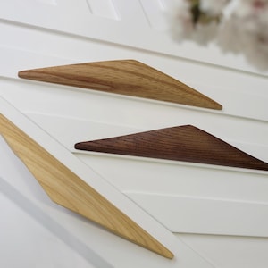 Könnte beinhalten: Drei Holzmöbelgriffe in verschiedenen Braun- und Beigetönen. Die dreieckigen Griffe sind auf einer weißen Oberfläche montiert. Die Holzmaserung ist sichtbar. Dekorative Hardware.