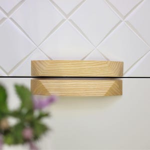 Puede incluir: Primer plano de un tirador de armario de madera clara con un diseño geométrico. El tirador es rectangular con bordes biselados. El fondo presenta una pared blanca texturizada con un patrón de diamantes.