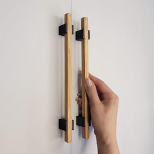 May include: Two wooden cabinet handles with a natural wood grain finish and black square mounting brackets. The handles are rectangular and vertically oriented, mounted on a white cabinet door. A hand is reaching for one of the handles.