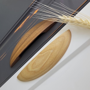 May include: Two wooden cabinet handles in different shades of brown and tan are displayed against a gray and white background. A sprig of wheat adds a natural element to the composition. The handles have a curved, ergonomic design.