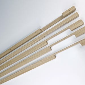 Peut inclure: Ensemble de poignées et de tirettes de meubles en bois non fini, disposées en diagonale sur fond blanc. Les poignées sont rectangulaires avec une découpe pour la prise, et le bois est de couleur brun clair naturel.