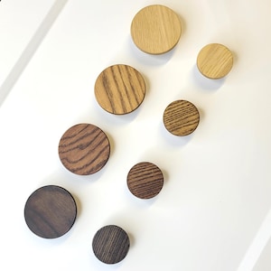 May include: A collection of eight wooden cabinet knobs in varying sizes and wood tones, including light oak, medium brown, and dark walnut. The knobs are arranged on a white surface, showcasing their natural grain patterns.