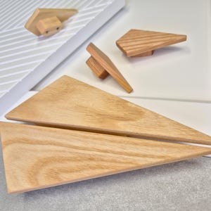 May include: A set of wooden cabinet hardware, including triangular drawer pulls and knobs. The light-coloured wood displays a natural grain. The set provides several pieces for a complete cabinet upgrade, suitable for various furniture styles.