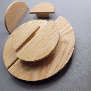 Könnte beinhalten: Eine Sammlung hellfarbiger Holzschrankknöpfe und Schubladengriffe. Die Knöpfe sind kreisförmig und halbkreisförmig, mit einer natürlichen Holzmaserung. Die Schubladengriffe haben eine rechteckige Basis und eine abgerundete Oberseite. Die Teile sind auf einer grauen Oberfläche angeordnet.