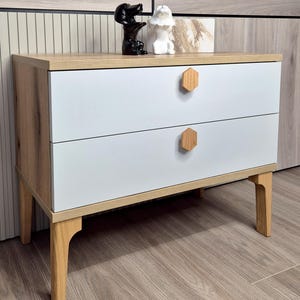 May include: A two-drawer nightstand with a light wood frame and white drawers. The drawers have hexagon-shaped wooden knobs. The nightstand is supported by four angled legs. Two decorative figurines sit on top.