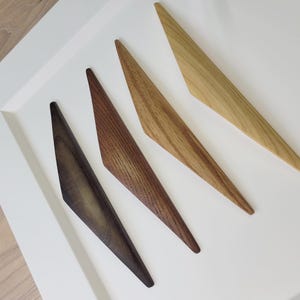 Puede incluir: Cuatro tiradores de madera para armarios en tonos marrones y beige, dispuestos diagonalmente sobre una superficie blanca. Los tiradores tienen un diseño angular único, con un extremo puntiagudo y una base más ancha. La veta de la madera es visible.
