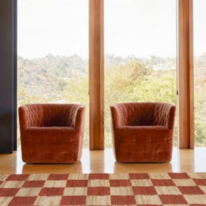 May include: Two rust-colored velvet armchairs with a tufted design are positioned in front of a large window. A checkered rug in red and beige tones is in the foreground. The chairs are on a wooden floor.
