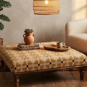 Può includere: Un pouf quadrato con un motivo geometrico giallo senape e nero, sostenuto da quattro gambe in legno scuro. La superficie superiore ospita un vaso in terracotta con una pianta grassa, due libri impilati e un vassoio in legno con una tazza in ceramica.