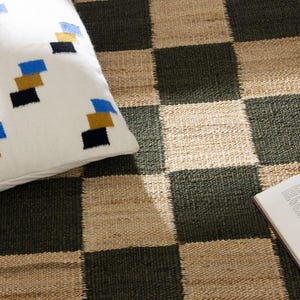 May include: A white pillow with a geometric pattern of blue, gold, and black squares rests on a checkerboard rug. The rug features alternating squares of dark green and natural beige. An open book is on the rug.
