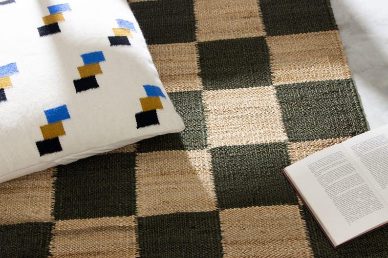 May include: A white pillow with a geometric pattern of blue, gold, and black squares rests on a checkerboard rug. The rug features alternating squares of dark green and natural beige. An open book is on the rug.