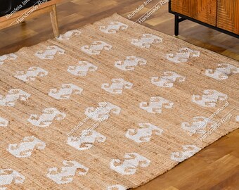 May include: A rectangular area rug featuring a natural tan woven fiber base with a repeating pattern of white, stylized, abstract shapes. The rug displays the text Bundela International Handmade Rugs printed across the surface in white.
