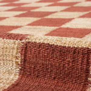 May include: Close-up of a woven rug with a repeating pattern of red and beige squares. The rug has a textured surface and a natural, earthy aesthetic. The design is a classic checkerboard.