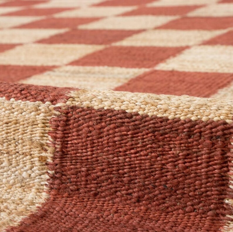 May include: Close-up of a woven rug with a repeating pattern of red and beige squares. The rug has a textured surface and a natural, earthy aesthetic. The design is a classic checkerboard.
