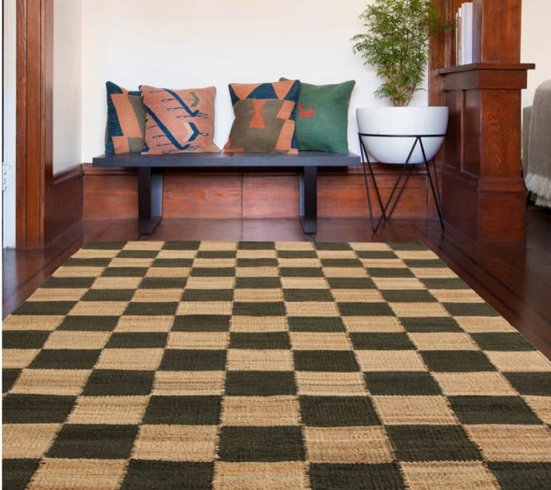 May include: A woven area rug with a checkerboard pattern in shades of green and tan. A black bench with decorative pillows in various colors sits against a wood-paneled wall. A white planter with a plant is also visible.