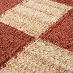 May include: Close-up of a woven rug with a geometric pattern. The rug features alternating squares of reddish-brown and beige, creating a textured surface. The weave appears to be made of natural fibers.