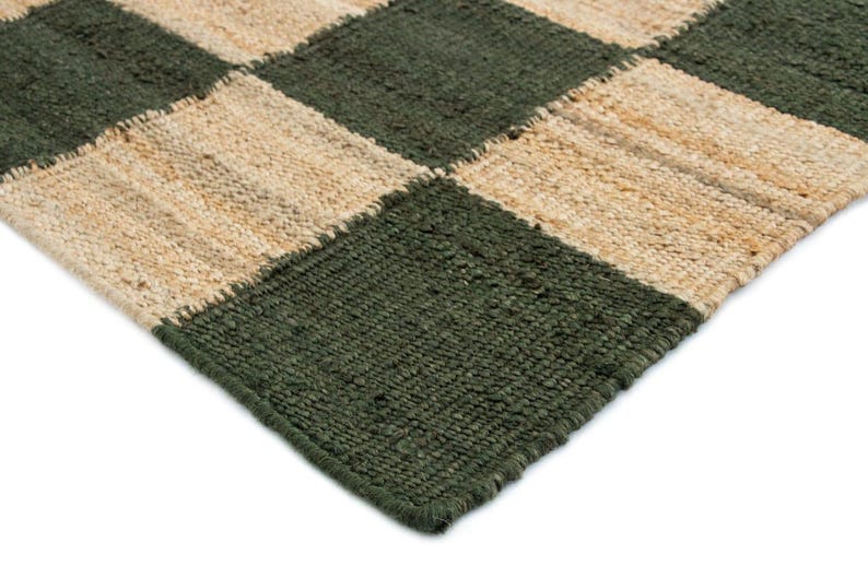 May include: Close-up of a woven rug with a checkerboard pattern. The rug features alternating squares of dark green and natural beige. The texture of the rug appears to be made of a natural fiber.