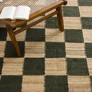 May include: A woven area rug with a checkerboard pattern in dark green and beige. A wooden chair with a woven seat holds an open book and a fringed throw. The rug's texture is visible.