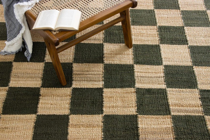 May include: A woven area rug with a checkerboard pattern in dark green and beige. A wooden chair with a woven seat holds an open book and a fringed throw. The rug's texture is visible.