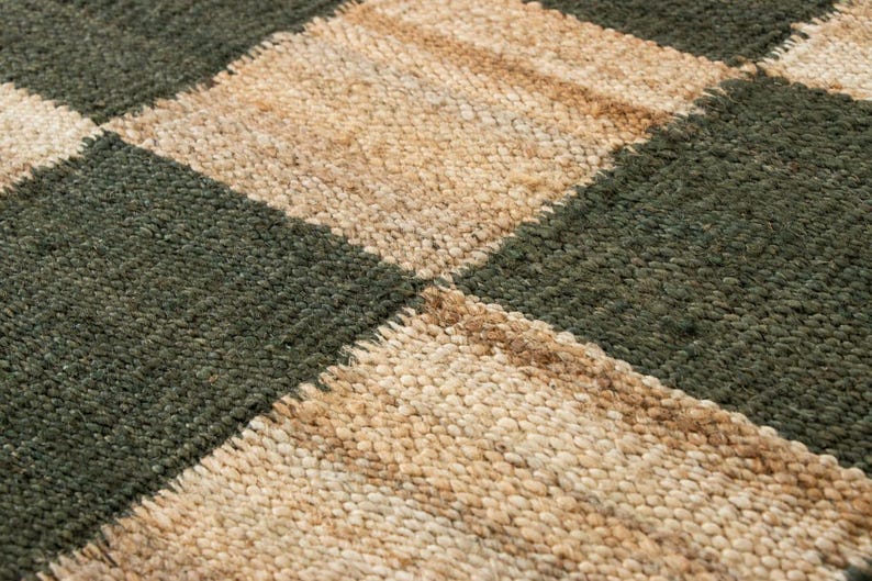 May include: Close-up of a woven rug with a checkerboard pattern. The squares alternate between a dark green and a natural beige color. The rug has a textured, nubby surface.