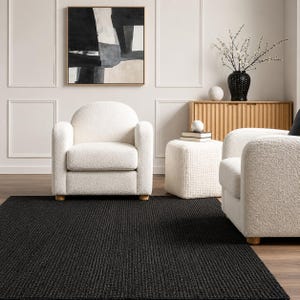 Puede incluir: Una alfombra rectangular negra con un diseño texturizado y acanalado. Dos sillones blancos y un otomán a juego están colocados sobre la alfombra. Un mueble de madera, obras de arte y objetos decorativos completan el ambiente del salón.