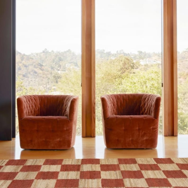 May include: Two rust-colored velvet armchairs with a tufted design are positioned in front of a large window. A checkered rug in red and beige tones is in the foreground. The chairs are on a wooden floor.