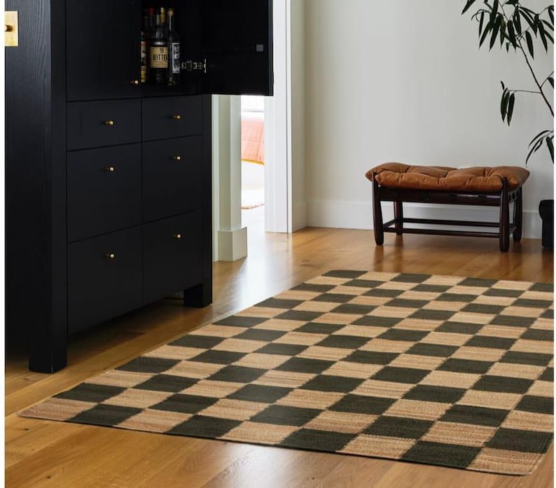 May include: A black and tan checkered rug on a hardwood floor. A black cabinet with gold knobs is on the left. A brown bench with a tufted cushion is in the background. A plant is on the right.