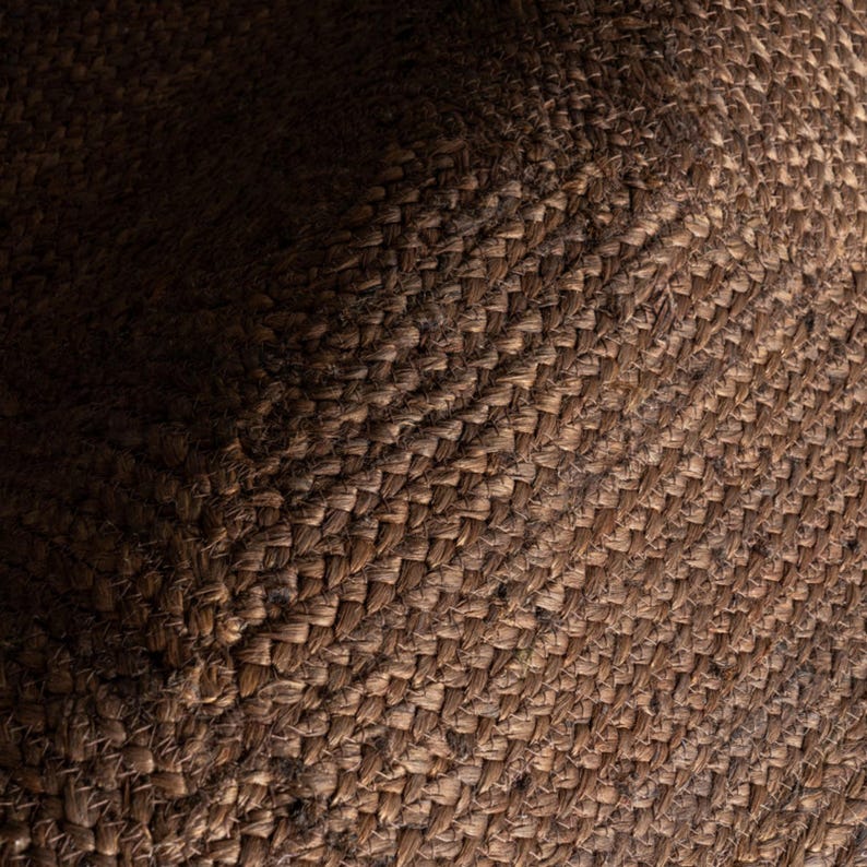 Może przedstawiać: Zbliżenie tkanego brązowego materiału o teksturowanej powierzchni. Materiał wydaje się być wykonany z naturalnych wł&oacute;kien, z diagonalnym wzorem utworzonym przez splot. Oświetlenie tworzy cienie i podkreśla fakturę.