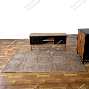 May include: A rectangular area rug featuring a natural tan woven texture with a repeating square geometric pattern. The rug is positioned on a wooden floor in front of a low wooden media console with black cabinet doors and an open center shelf.