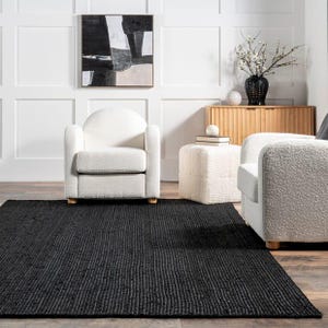 Op de afbeelding: Een zwart rechthoekig tapijt met een geribbelde textuur is het middelpunt van een woonkamer. Twee witte fauteuils en een kleine poef zijn rond het tapijt geplaatst. Een houten kast en abstracte kunst maken de inrichting compleet.