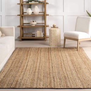 May include: A rectangular, natural-toned jute rug with a ribbed texture. The rug is placed on a light-colored floor, and is surrounded by neutral-toned furniture, including a sofa, chair, and shelving unit. The rug's dimensions are approximately 8 x 10 feet.