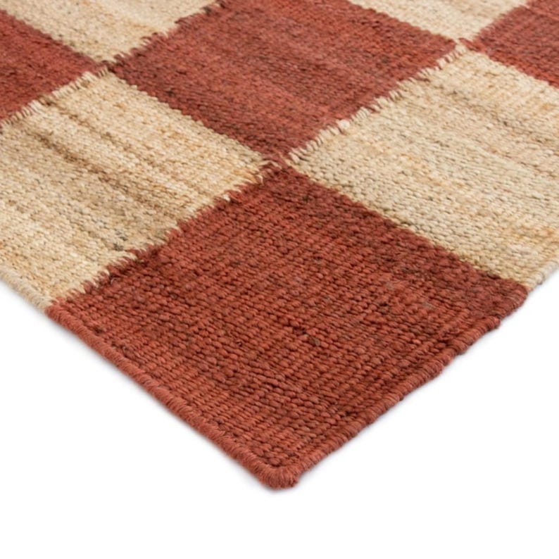 May include: Close-up of a woven rug with a checkerboard pattern. The rug features alternating squares of reddish-brown and beige. The texture of the rug appears coarse and natural, suggesting a jute or similar material. The rug's edge is visible.