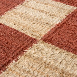 May include: Close-up of a woven rug with a geometric pattern. The rug features alternating bands of reddish-brown and beige, creating a textured, rectangular design. The fibers appear thick and natural, suggesting a handmade or artisanal quality.