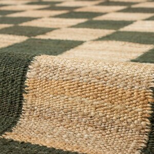 May include: A close-up of a woven rug with a checkerboard pattern. The rug features alternating squares of dark green and natural beige. The texture of the rug is visible, with a rolled-up edge showing the thickness and construction.