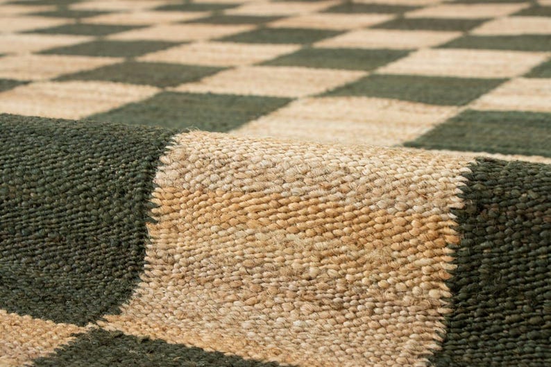 May include: A close-up of a woven rug with a checkerboard pattern. The rug features alternating squares of dark green and natural beige. The texture of the rug is visible, with a rolled-up edge showing the thickness and construction.