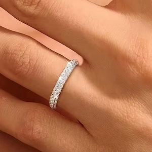 Puede incluir: Un anillo plateado con una banda completamente incrustada con pequeñas piedras claras y brillantes. El anillo se lleva en un dedo, mostrando su delicado diseño y su brillo brillante. El anillo es una pieza de joyería clásica.