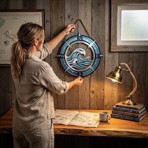 May include: A circular metal wall art piece featuring a stylized ocean wave design in shades of blue and white, framed by a compass rose with cardinal directions N, S, E, and W. The piece is suspended by a thick jute rope against a wood wall.