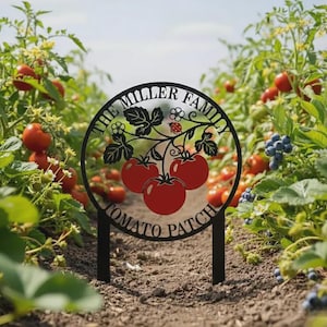 May include: A circular black metal garden sign featuring a cutout design of three red tomatoes on a vine with leaves and a small ladybug. The text "THE MILLER FAMILY" and "TOMATO PATCH" is engraved along the top and bottom of the circular frame.