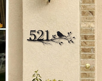 Puede incluir: Letrero de número de casa de metal negro con el número 521, un pájaro y ramas con hojas. El letrero está montado en una pared de estuco beige, con una lámpara encima y una columna de ladrillos a la derecha.