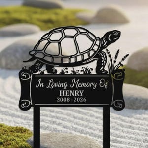 May include: A black metal memorial garden stake featuring a turtle design. The stake reads "In Loving Memory Of HENRY 2008-2026". The turtle is perched atop a decorative plaque with scrollwork and grass accents.