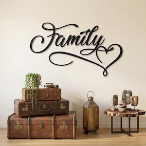 May include: A black metal "Family" sign in cursive script with a heart detail, mounted on a white wall. The sign is above a stack of vintage brown suitcases, a lantern, and a small wooden table with decorative objects.