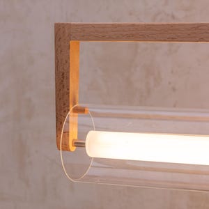 May include: Modern wall sconce featuring a rectangular wooden frame and a clear glass tube. The tube houses a bright, cylindrical light source. The fixture is mounted on a textured, neutral-toned wall.