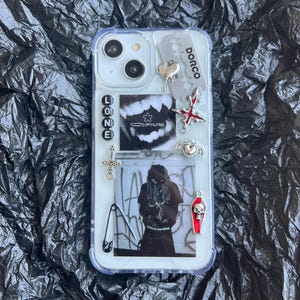 Peut inclure: Une coque de téléphone transparente ornée de breloques variées dont une lame de rasoir Dorco argentée, un cœur, une croix, une épingle à nourrice et un cercueil avec un crâne. Elle présente un graphique noir et blanc de dents, le mot LONE et une photo.